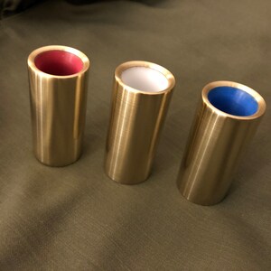Op de afbeelding: Drie gouden metalen slide-gitaar-slides met verschillende gekleurde inzetstukken. De slide links heeft een rode inzet, de middelste slide heeft een witte inzet en de slide rechts heeft een blauwe inzet.