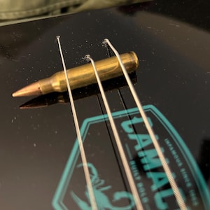 Puede incluir: Primer plano de un ukelele negro con cuatro cuerdas y una bala dorada descansando sobre las cuerdas. El ukelele tiene un logotipo de color turquesa que dice "TAMAL" con las palabras "BUILT SOLID" e "INSPIRED SINCE 1963" debajo.