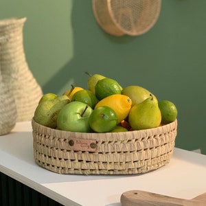 May include: A woven wicker basket filled with green pears, limes, and yellow lemons. The basket has a brown leather tag that says "Bohemian Living".