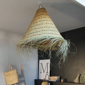 May include: A bedroom with a large bed featuring white linen sheets and grey bedding. The bed has a black headboard and a large woven lampshade hanging above it. There are two white pillows with pom-poms on the bed. The room has a black door and white walls.