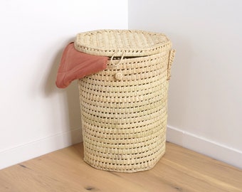 May include: A natural woven wicker laundry hamper with a lid and handles. The hamper is in a corner of a room with a white wall and wood floor. A piece of red fabric is hanging out of the hamper.