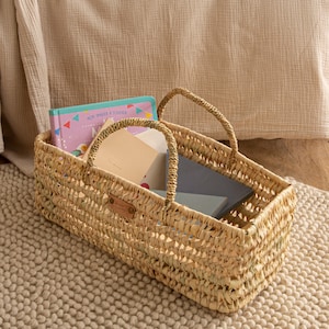 Puede incluir: Una cesta rectangular de mimbre trenzado con asas de cuero. La cesta está llena de libros, incluyendo uno con el título "Mon Imaginaire a Toucher".