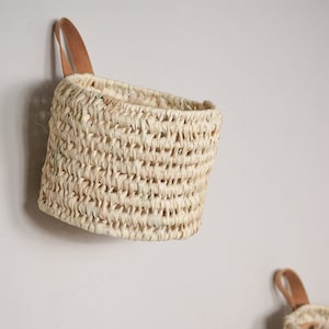 Op de afbeelding: Een natuurlijke geweven wandmand met een leren handvat. De mand is gemaakt van licht beige materiaal en heeft een afgeronde vorm.