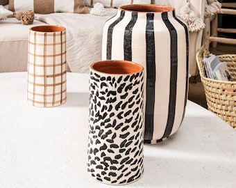 May include: Three ceramic vases with different patterns. The tallest vase is white with black vertical stripes. The middle vase is white with black abstract spots. The shortest vase is white with brown plaid.