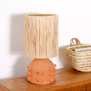 Peut inclure: Une lampe de table en terre cuite avec un abat-jour cylindrique en raphia. La base est de forme arrondie avec de petites protubérances arrondies. La lampe est posée sur une surface en bois, avec un panier tressé en arrière-plan.