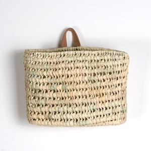 Peut inclure: Un panier rectangulaire tressé en fibres naturelles de couleur beige clair avec une texture à mailles ouvertes visible. Le panier comporte une unique poignée en cuir arquée et centrée sur le bord supérieur pour une suspension murale.