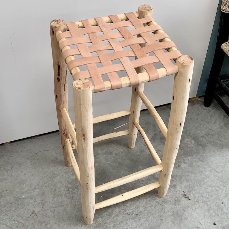 May include: A handcrafted wooden stool with a woven leather seat. The light-colored wood frame features a natural, unfinished look, with the seat made of interwoven tan leather strips. The stool has four sturdy legs and is designed for seating.