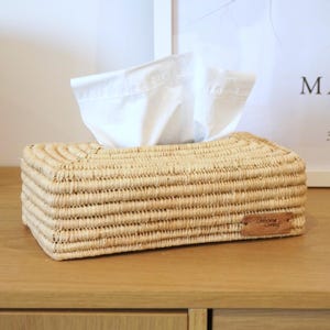 Peut inclure: Un distributeur de mouchoirs rectangulaire en fibres naturelles tressées, avec une petite étiquette en cuir marron portant l'inscription "Belong Living". Des mouchoirs blancs dépassent du dessus. Le tout est posé sur une surface en bois.