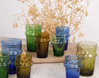 May include: A collection of eight Moroccan tea glasses in various shades of blue, green, and brown. The glasses are arranged on a wooden surface with dried flowers in the background.