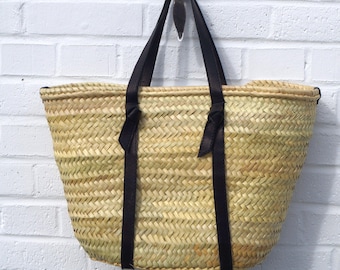 May include: A woven straw tote bag with black leather handles and accents, hanging against a white brick wall. The bag has a rectangular shape and is designed for carrying items.