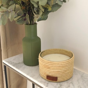 Puede incluir: Una vela grande, redonda y tejida con un interior de cera blanca. La vela está sobre una mesa de mármol blanco junto a un jarrón verde con vegetación.