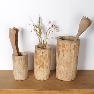 Peut inclure: Trois vases en bois avec une finition en écorce naturelle, l'un contenant des fleurs séchées, l'autre des ustensiles en bois et le troisième vide. Les vases sont disposés sur une surface en bois.