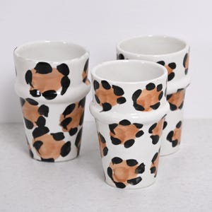 Peut inclure: Trois tasses en céramique blanche avec un motif léopard distinctif. Les tasses présentent un motif répété de taches marron et noires, créant une esthétique audacieuse et élégante. Les tasses ont une forme cylindrique avec un bord légèrement évasé.