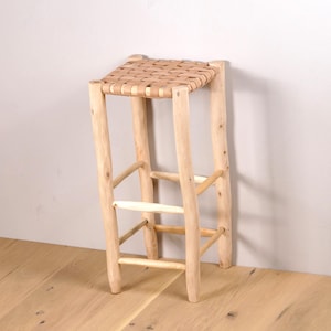 May include: A light-colored wooden stool with a woven seat. The stool has four legs and a rectangular seat made of woven strips. The legs are connected by horizontal and angled supports, creating a sturdy structure. The stool is made of natural wood.