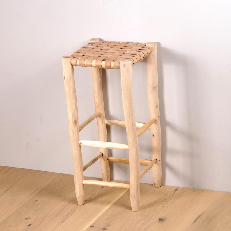 May include: A light-colored wooden stool with a woven seat. The stool has four legs and a rectangular seat made of woven strips. The legs are connected by horizontal and angled supports, creating a sturdy structure. The stool is made of natural wood.