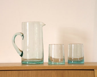 May include: A set of three clear glass containers, including a pitcher and two tumblers. The pitcher has a handle and a rounded base. The tumblers are cylindrical and have a slightly flared rim.