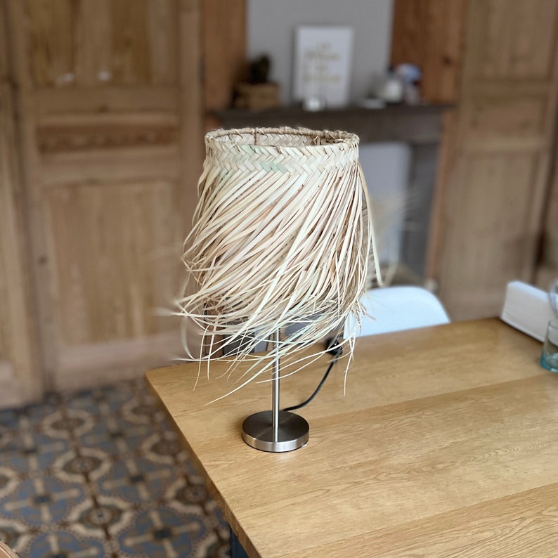 May include: A table lamp with a natural woven shade made of straw. The shade is fringed and has a natural, light brown color. The lamp base is made of silver metal.
