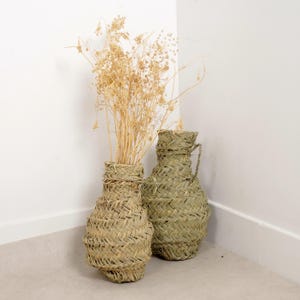 May include: Two woven vases, one with dried beige flowers, stand in a corner. The vases are made of a light green material with a textured, basket-like weave. One vase has a handle.
