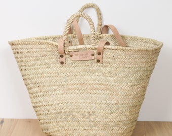 Puede incluir: Un bolso tote de paja tejida con dos asas y correas de cuero. El bolso tiene forma rectangular con una abertura ancha. El texto "Bohome Living" está en una etiqueta de cuero adjunta al bolso.