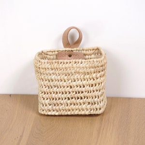 Peut inclure: Un petit panier mural rectangulaire en paille tressée avec une anse en cuir. Le panier porte une étiquette "Boheme Living".