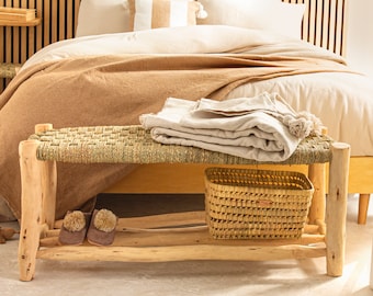 May include: A woven wood bench with a natural woven seat and a lower shelf. The bench has a woven basket on the shelf and two fuzzy slippers on the lower shelf. The bench is in front of a bed with a brown blanket and white pillows.