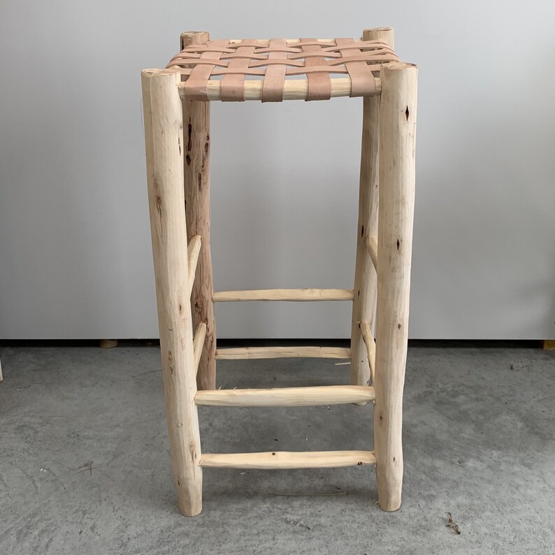 May include: A tall, rustic bar stool with a woven leather seat. The frame is constructed from light-colored, unfinished wood, featuring natural knots and textures. The stool has a ladder-style footrest and is designed for a bar or counter setting.