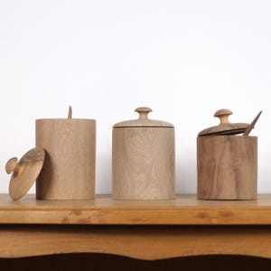 Può includere: Tre contenitori in legno con coperchi. I contenitori sono cilindrici e hanno una finitura in legno naturale. I coperchi sono anch'essi in legno e hanno un pomo in cima. I contenitori sono disposti in fila su una superficie in legno.