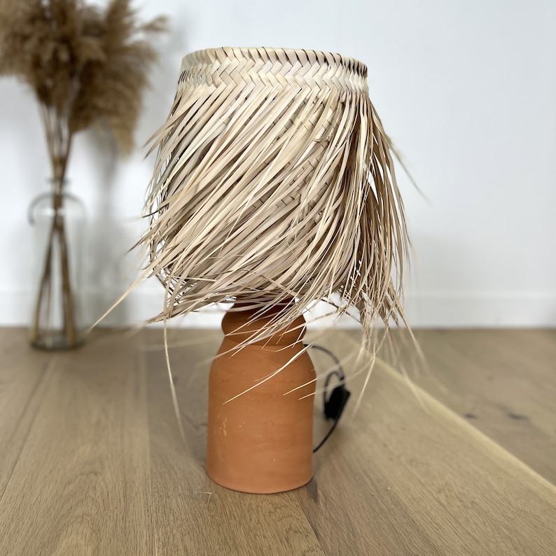 May include: A terracotta table lamp with a woven natural fiber shade. The shade has a fringed edge and is made of natural materials.