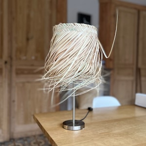 May include: A table lamp with a natural woven shade made of straw. The shade is a light brown color and has a frayed edge. The lamp base is silver metal.
