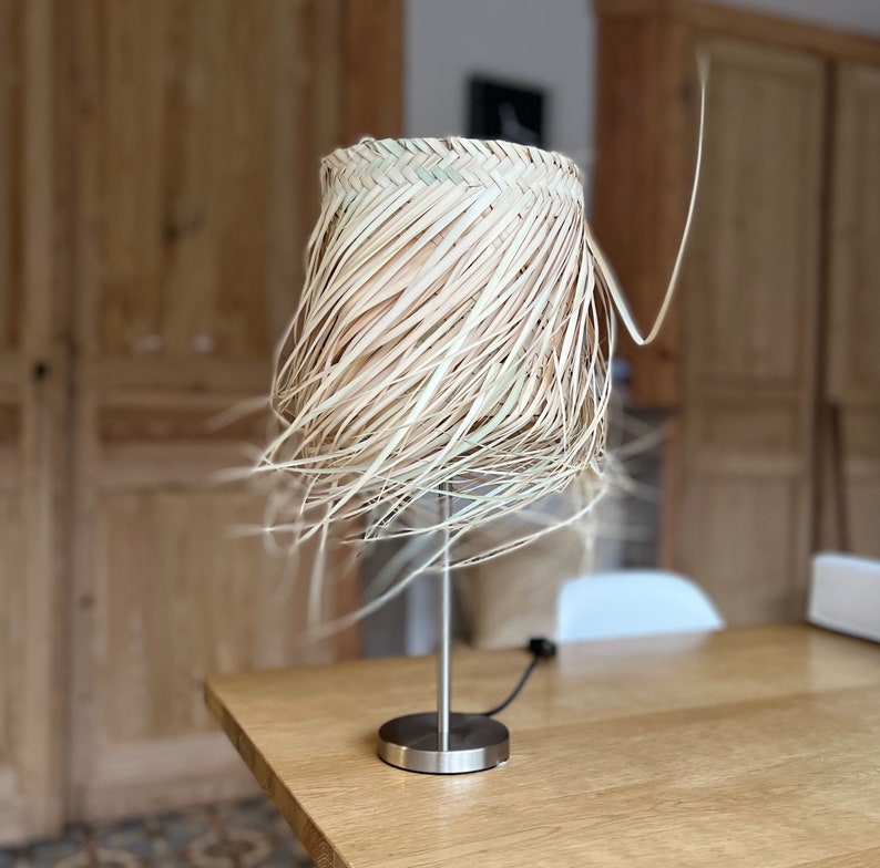 May include: A table lamp with a natural woven shade made of straw. The shade is a light brown color and has a frayed edge. The lamp base is silver metal.
