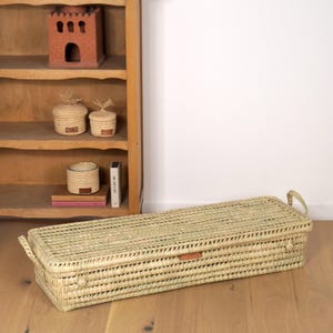 Puede incluir: Una cesta rectangular tejida con tapa y asas, hecha de fibras naturales. La cesta está colocada sobre un suelo de madera. Otros objetos decorativos están en una estantería de madera en el fondo. Mide aproximadamente 100 cm de largo.