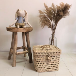 Puede incluir: Una cesta de almacenamiento tejida con tapa y asa. La cesta es de color marrón claro y tiene un patrón tejido. La cesta está sentada en un suelo de madera junto a un taburete de madera y un jarrón de hierba de la pampa seca.