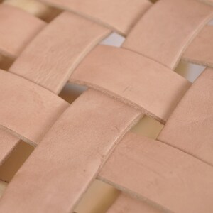 May include: Close-up of a woven leather pattern. The leather strips are a light tan color, creating a basket-weave design. The texture of the leather is visible, with slight variations in color and tone.