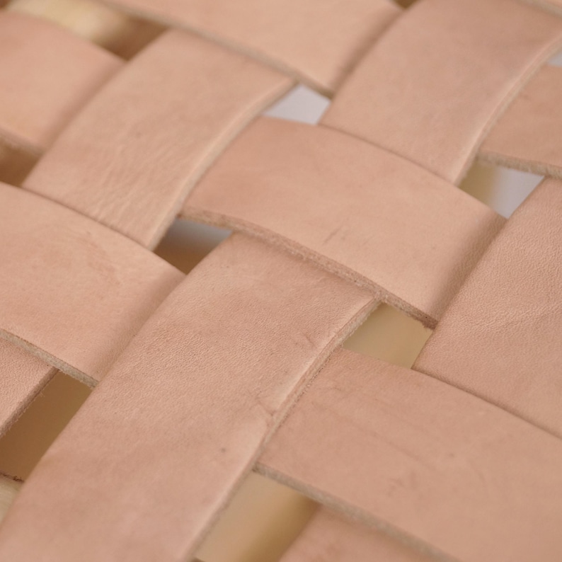 May include: Close-up of a woven leather pattern. The leather strips are a light tan color, creating a basket-weave design. The texture of the leather is visible, with slight variations in color and tone.