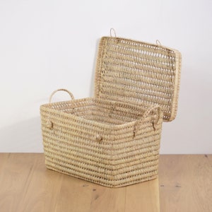 Peut inclure: Un panier de rangement en osier naturel avec un couvercle. Le panier est fait d'un mat&eacute;riau brun clair et a une forme rectangulaire. Le couvercle est ouvert et le panier est pos&eacute; sur un sol en bois.