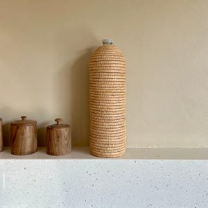 May include: A tall, cylindrical water bottle encased in woven tan material. The bottle has a clear plastic top. Two small wooden containers with lids are to the left of the bottle, all on a white surface.