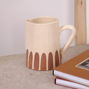 Puede incluir: Jarra de cerámica color crema con asa curva. La base de la jarra está decorada con formas ovaladas marrones verticales. La jarra está sobre una superficie gris junto a una pila de libros.