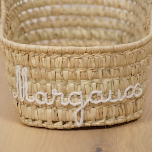 Può includere: Un cesto intrecciato naturale con una scritta in crochet bianco che dice "Margaux".