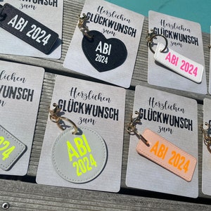 May include: A set of eight keychains with different colored backgrounds and text that says "Herzlichen Gl&uuml;ckwunsch zum ABI 2024".