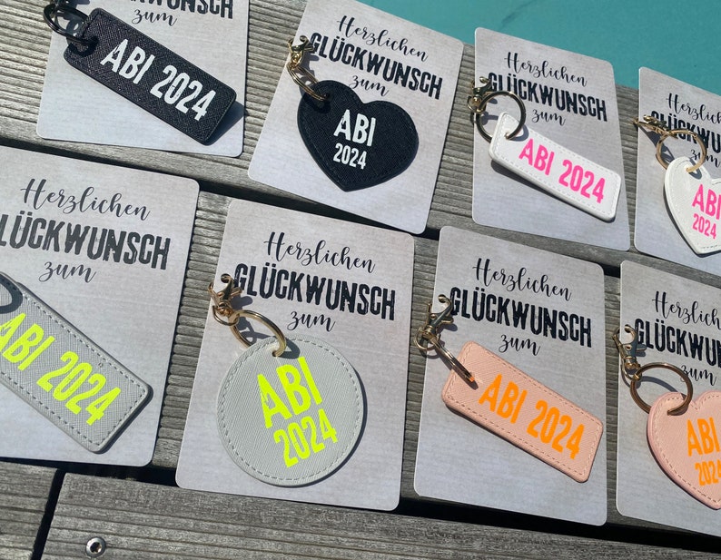 May include: A set of eight keychains with different colored backgrounds and text that says "Herzlichen Gl&uuml;ckwunsch zum ABI 2024".