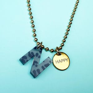 Könnte beinhalten: Eine goldfarbene Kugelkette mit einem Anhänger in Form des Buchstaben "Z" mit blau-grauem Muster und einem kleinen goldenen Scheibenanhänger mit der Aufschrift "HAPPY". Die Halskette wird vor einem hellblauen Hintergrund präsentiert.