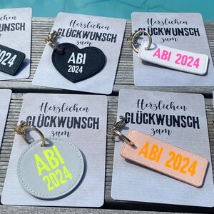May include: A set of six keychains with different designs. Each keychain features a black, white, gray, or pink leather tag with the text "ABI 2024" in a contrasting color. The keychains are displayed on white cards with the text "Herzlichen Gl&uuml;ckwunsch zum" in black.