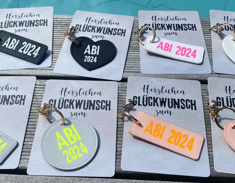 May include: A set of six keychains with different designs. Each keychain features a black, white, gray, or pink leather tag with the text "ABI 2024" in a contrasting color. The keychains are displayed on white cards with the text "Herzlichen Gl&uuml;ckwunsch zum" in black.