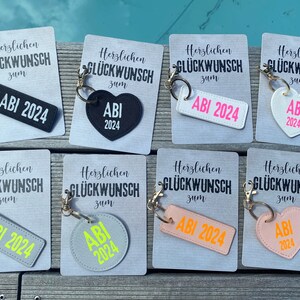 May include: Eight different keychains with the text "Herzlichen Gl&uuml;ckwunsch zum ABI 2024" on them. The keychains are in different shapes and colors, including a rectangle, a heart, and a circle. The keychains are all made of leather.