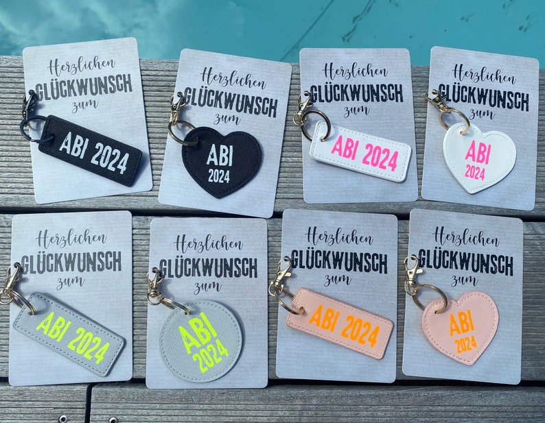 May include: Eight different keychains with the text "Herzlichen Gl&uuml;ckwunsch zum ABI 2024" on them. The keychains are in different shapes and colors, including a rectangle, a heart, and a circle. The keychains are all made of leather.