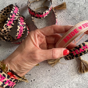 Könnte beinhalten: Armbänder mit Leopardenmuster und rosa Text, darunter "Bride Fifi" und "Team Bride". Jedes Armband hat eine goldene Quaste. Diese Armbänder sind ein lustiges Accessoire für einen Junggesellinnenabschied.
