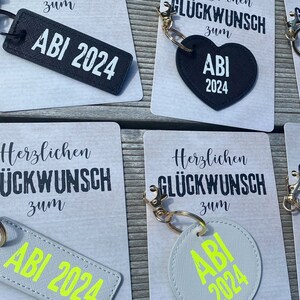 May include: A set of five keychains with different designs. Each keychain features a black and white card with the text "Herzlichen Gl&uuml;ckwunsch zum" and a black or gray keychain with the text "ABI 2024". The keychains are perfect for celebrating a graduation.
