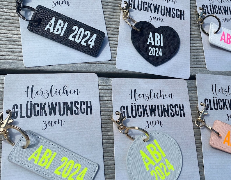 May include: A set of five keychains with different designs. Each keychain features a black and white card with the text "Herzlichen Gl&uuml;ckwunsch zum" and a black or gray keychain with the text "ABI 2024". The keychains are perfect for celebrating a graduation.