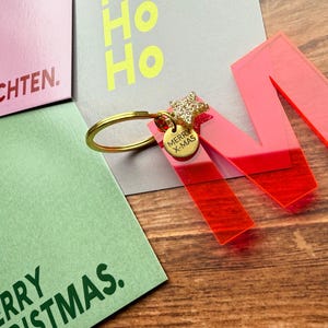 Puede incluir: Un llavero con la letra "M" de acrílico rojo brillante, con un anillo dorado y un charm "Merry X-Mas". El llavero está junto a tarjetas navideñas con texto en inglés y alemán. El fondo es una superficie de madera.