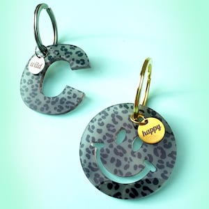 Könnte beinhalten: Zwei Schlüsselanhänger mit grauem Leopardenmuster. Einer ist ein Buchstabe "C" mit dem Wort "wild" auf einem kleinen silbernen Anhänger. Der andere ist ein Smiley mit dem Wort "happy" auf einem goldenen Anhänger. Beide haben silberne und goldene Schlüsselringe.